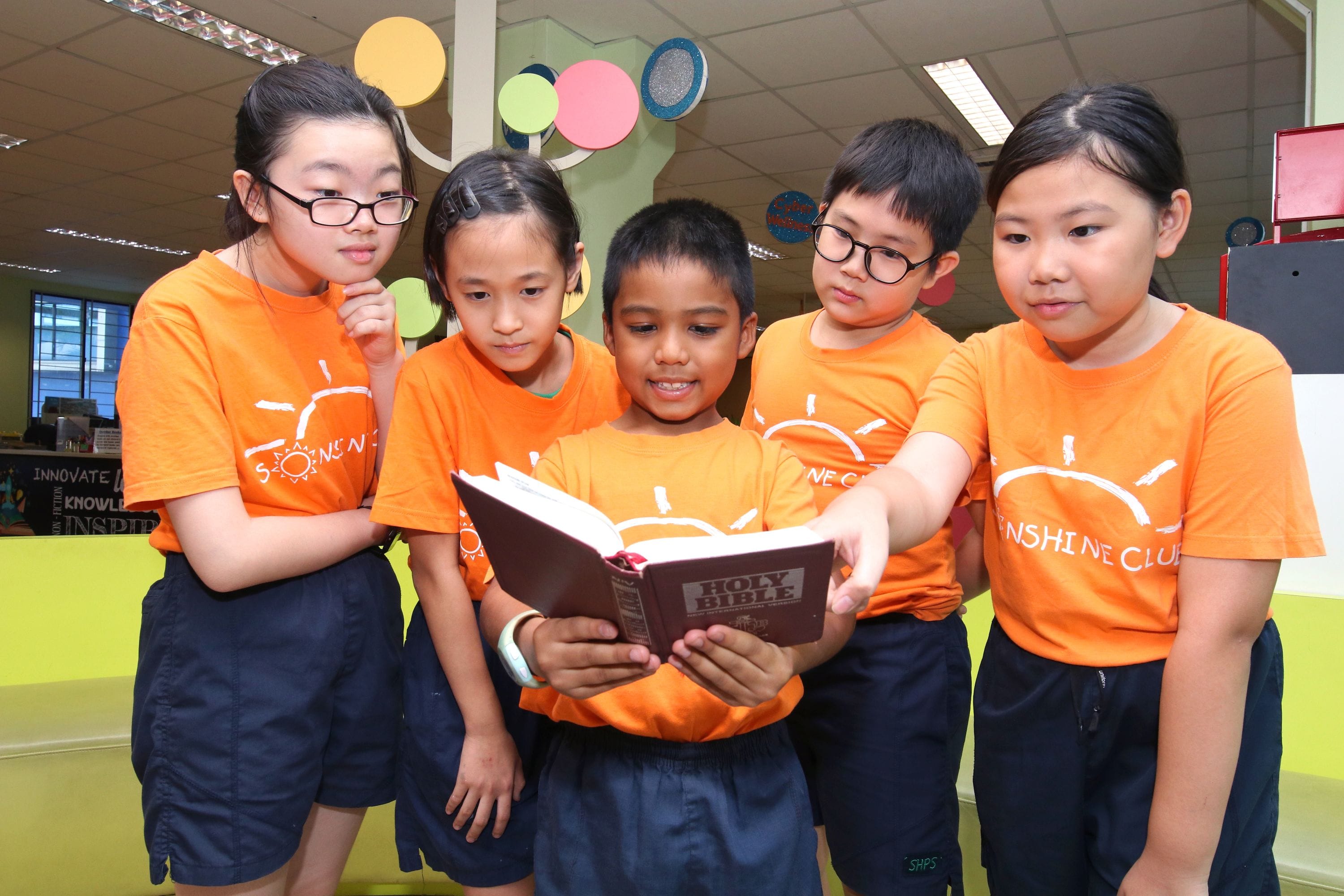 Five children in orange Sunshine Club shirts gather around a boy holding an open Holy Bible indoors.